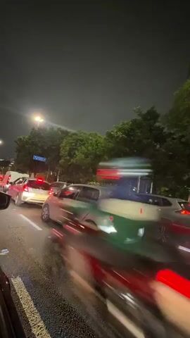 The video depicts a traffic jam at night. Several cars are stationary on a multi-lane road, with their taillights illuminated. A silver hatchback is centrally positioned in the frame, with a silver SUV to its right and a red car to its left. Streetlights illuminate the scene, and trees line the roadside in the background. The sky is dark, suggesting it is late at night. The traffic appears congested with multiple vehicles clustered together. The overall impression is of a typical urban traffic situation at nighttime.