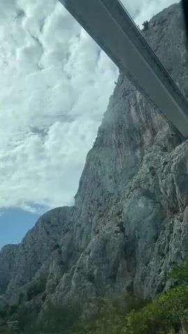 The video begins with a view of a large rock face with clouds in the sky. The perspective then shifts to the interior of a vehicle, showing a blurry view of the road and rock walls as it enters a tunnel. The camera pans slightly, revealing the rough texture of the rock walls. The video ends with the vehicle continuing through the dimly lit tunnel.