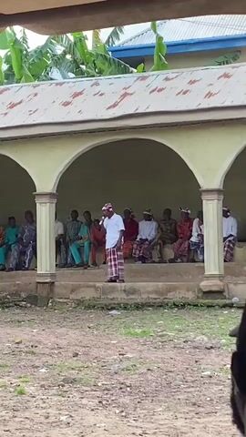 This video captures an outdoor gathering with a group of people seated under a covered structure with arches. A man stands in front of them, speaking into a microphone. He is wearing a white shirt and a red and white checkered garment around his waist, and a patterned cap. The seated individuals are dressed in various traditional African attire, some with headwraps.

The setting appears to be a community space, possibly a gathering hall or a ceremonial area, with a dirt ground in the foreground and a building with a metallic roof and banana trees visible in the background. The lighting suggests it is daytime, likely morning or afternoon, with soft shadows indicating diffused sunlight. The overall atmosphere seems to be one of a formal or semi-formal assembly. The people appear attentive to the speaker, suggesting an event of importance such as a speech, announcement, or performance. No text is visible in the video. The location context of Nguzu Edda, Nigeria, places this scene within a specific cultural and geographical setting in West Africa.