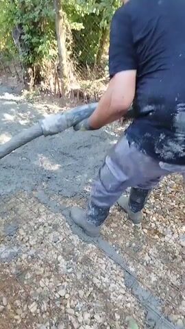 A man in work clothes and boots is operating a concrete pump hose. He is standing in an outdoor area with trees and foliage in the background. The hose is directing wet concrete onto a surface that appears to be a mix of gravel and reinforcement mesh. The man is holding the hose with gloved hands and appears to be moving it to distribute the concrete. The concrete is flowing from the end of the curved hose onto the ground.