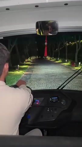 The video shows a first-person perspective from inside a vehicle, likely a shuttle bus or large golf cart, at night. A man, seen from behind, is driving the vehicle. He wears a light-colored long-sleeved shirt and is focused on the road ahead, with his hands on the steering wheel. His reflection is visible in the rearview mirror, which also has a red decorative tassel hanging from it.

The vehicle is traveling along a paved, possibly cobblestone or brick, path. Both sides of the path are lined with trees and numerous small, ground-level lights that illuminate the way, creating a well-lit corridor through the darkness. In the distance, two structures or gates are visible, also illuminated. The setting suggests a resort, hotel, or private estate, especially given the decorative lighting and curated landscape. The time of day is clearly night. No specific text is legible on the vehicle's dashboard. The location is Mu Cang Chai, Vietnam.