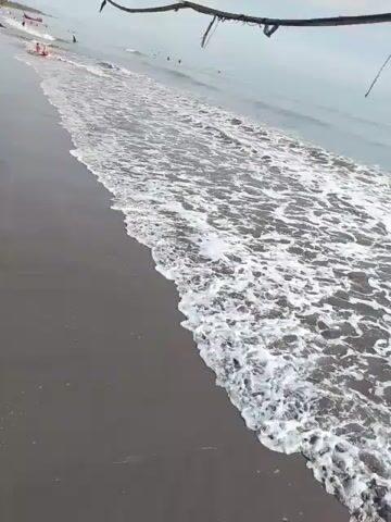 The video shows a view of a beach with gentle waves lapping at the shore. The sand appears dark and wet. In the distance, several people can be seen on the beach and in the water, and a few small boats are visible. The sky is overcast. The camera is positioned slightly above the beach, looking down towards the water. The overall atmosphere is calm and serene.