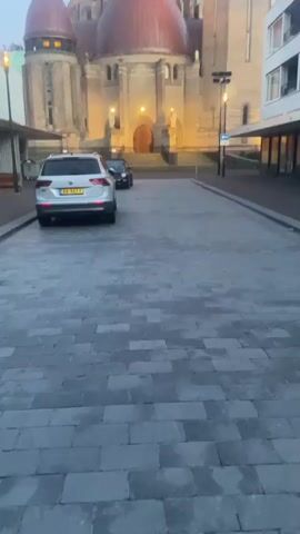 The video opens showing a white SUV parked on a cobblestone street. In the background, a large building with multiple domes is visible. As the video progresses, the camera pans upwards, revealing the full facade of the building, which appears to be a church or cathedral. The camera then focuses on a tall, slender tower attached to the building, which has a clock face near the top. The sky is a clear blue. The video is shot from a low angle, looking up at the architecture.