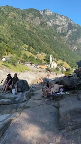 A group of people are enjoying a summer day by a river in Verzasca, Switzerland. The scene is dominated by a steep, forested mountainside with scattered houses and a prominent church with a bell tower. In the foreground, a rocky riverbed features several individuals sunbathing and relaxing on large, smooth stones. Some are sitting together, chatting and enjoying the scenery, while others are positioned slightly apart, lost in their own thoughts or perhaps observing the activities in the water.

The time of day appears to be late afternoon, as indicated by the warm, golden light casting shadows across the rocks and highlighting the lush greenery of the mountains. The weather is clear and sunny, with a bright blue sky visible above the peaks. The overall atmosphere is one of leisure and relaxation, with the natural beauty of the Verzasca Valley providing a picturesque backdrop for a peaceful summer outing. The subjects are dressed in casual summer attire, including swimwear, further emphasizing the warm weather and relaxed mood. No visible text is present in the media file.