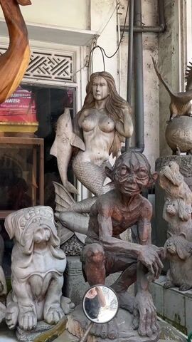 The video shows several wood sculptures, including a mermaid, Gollum, and a dog. The mermaid is holding a dolphin and has long hair. Gollum is crouching down and has a sinister smile. The dog is wearing a hat. The sculptures are all displayed outside of a building.