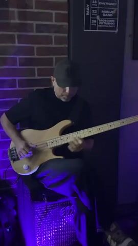 A man in a dark-colored cap, dark t-shirt, and blue pants is playing a bass guitar while sitting on a black amplifier. He sits in front of a brick wall in a dimly lit space. A second man, also wearing a dark-colored cap, stands next to a small black table and operates a laptop with a lighted keyboard. The space is illuminated with purple lighting. Behind the men, a chalkboard lists items and prices. A May events calendar is visible on the wall.