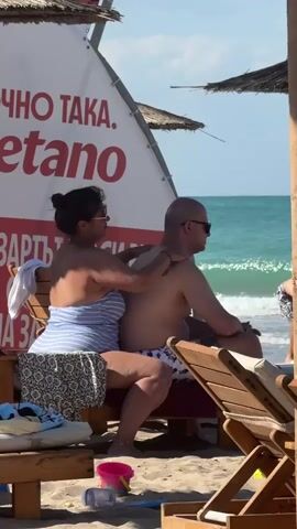 A woman is giving a man a shoulder massage on a beach. They are both sitting in lounge chairs. The man is facing away from the camera, and the woman is sitting behind him, with her hands on his shoulders. There is a large banner in the background with text on it. The sea is visible behind them, with waves breaking on the shore. Other beachgoers are visible in the distance. There are beach umbrellas and more lounge chairs in the foreground.