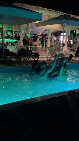 A group of young adults are socializing in a swimming pool at night. The pool is illuminated with bright blue lights. Behind the pool, there is a bar area with people gathered and a few lounge chairs. The overall atmosphere appears to be a lively outdoor party or gathering.