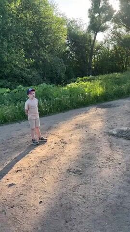 A young boy in a blue and red baseball cap, beige t-shirt, and beige shorts stands on a dirt path in a sunny, tree-lined area. He kicks a deflated soccer ball towards the camera, and it bounces on the path before rolling back to him. The boy looks at the camera and then kicks the ball again, this time bouncing it off a low invisible object and catching it with his feet. He looks towards the camera as the video ends.