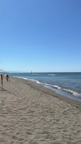 The video opens with a view of a sandy beach and the ocean under a clear blue sky. In the distance, a sailboat is visible on the water. As the video progresses, the camera pans slightly to the right. A buouy line with red and white markers appears in the water. Then, a person in a bikini is seen standing in the shallow water, facing away from the camera. The waves gently lap at the shore. The scene is peaceful and summery.