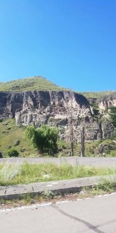 A vehicle is seen driving on a road. The vehicle passes by a steep, rocky cliff face with visible caves or openings carved into it. There are also trees and green vegetation present in the landscape. The sky is clear and blue.