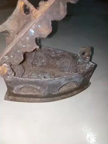 The video features a close-up, panning view of an antique, heavily rusted cast iron flat iron. The iron exhibits significant rust and corrosion on its metal components, including the base, the upper plate, and the handle support. Patches of blue-green discoloration, consistent with verdigris, are visible on the metal surfaces. The handle is made of dark, weathered wood. The iron rests on a light-colored, flat surface, possibly a tile floor, with a dark, out-of-focus background. The camera slowly rotates around the stationary object, showcasing its aged condition from various angles.