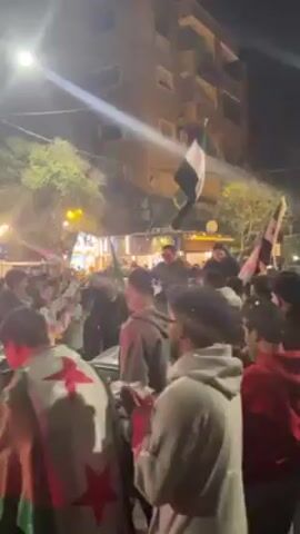 The video shows a large crowd of people celebrating outdoors at night. The crowd is tightly packed and the atmosphere appears lively. Many people are waving Syrian flags, which are visible throughout the crowd. Some individuals are wearing clothing adorned with the Syrian flag as well. The celebrations are taking place on a city street illuminated by streetlights. Buildings can be seen in the background, adding to the urban environment of the scene. The overall impression is one of collective joy and enthusiasm, likely in response to an event or occasion related to Syria.