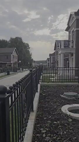 The video shows a quiet residential street lined with brick houses. A black metal fence runs along the right side of the frame, separating the sidewalk from the yards of the houses. The sky is overcast with gray clouds, and the overall lighting suggests it is daytime, possibly early morning or late afternoon. The street appears to be paved, and there are trees on the left side of the street. The houses have a similar architectural style, with visible balconies and decorative elements. There are no people or vehicles visible in the frame, suggesting a peaceful and undisturbed environment. The video captures a still, somewhat somber, atmosphere.