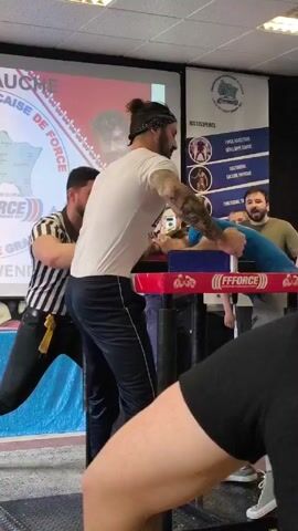 Here's a description of the video:

The video shows an arm-wrestling match taking place in what appears to be a gymnasium or large meeting room. Two men are positioned at an arm-wrestling table, with their elbows resting on the pads and hands gripping each other. One man wears a white t-shirt and dark pants, with his hair in a top knot, and the other wears a blue t-shirt and gray pants. A referee in a black and white striped shirt stands between them, overseeing the match.

Behind the table, there is a projector screen displaying a graphic related to arm wrestling, with text in French. There are also people standing around watching the match. The overall setting appears to be a local or regional arm-wrestling competition.