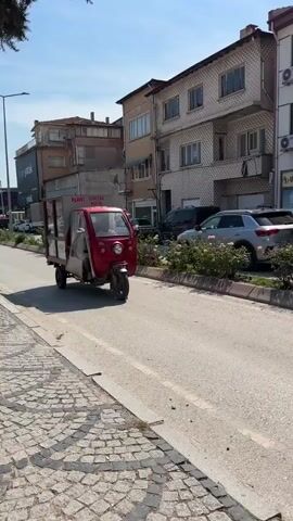 A red, three-wheeled motorized vehicle with a food stall attached to the back drives away from the camera on a sunny day. Two men are visible in the vehicle's cabin. The food stall is made of wood and glass and advertises "Nohutlu Pilav" and "Tencere Köfte." Buildings line both sides of the street. The sky is blue with scattered white clouds.