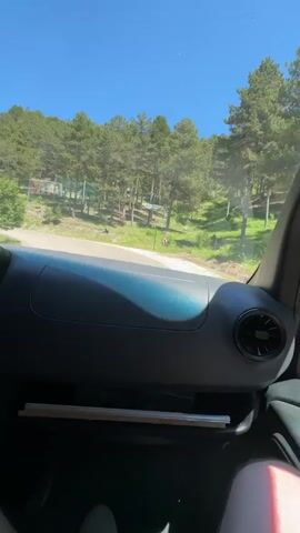 The video shows a view from inside a vehicle, looking out through the windshield. The dashboard of the vehicle is visible in the foreground, with a black panel and an air vent on the right side. A shelf below the panel holds some flat objects. Outside the vehicle, a paved road curves to the left, bordered by a grassy slope on the right and trees on the left. The trees are tall and green, with a clear blue sky above. The road appears to be in a rural or mountainous area. There are some colorful signs visible on the left side of the road, in the distance. The lighting suggests it is daytime, with bright sunlight.