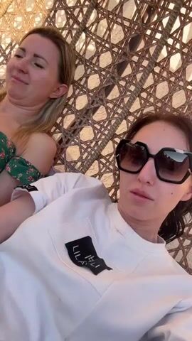 Two women are relaxing in a wicker chair. The woman in the foreground is wearing a white shirt and large sunglasses. The woman behind her is wearing a green floral top. The lighting suggests it is daytime. The background is a wicker texture.
