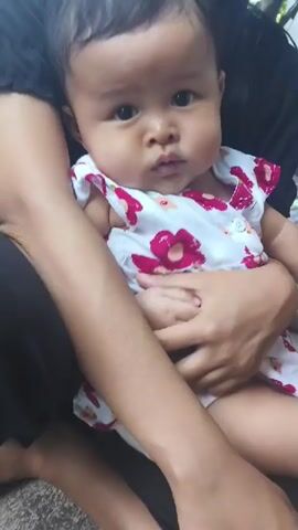 A baby is being held by an adult. The baby is wearing a white dress with pink flowers. The baby is looking at the camera. The adult is wearing a black shirt. The background is blurry but appears to be outdoors.