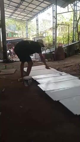 A person wearing a black t-shirt, dark shorts, and a hat is painting a long row of white rectangular panels laid out on the ground. The person is using a paint roller and a paint can, and appears to be working under a covered structure with a metal roof supported by metal beams. The environment is outdoors or in an open-air workshop, with dirt ground and vegetation visible in the background. The panels are arranged in a line, creating a path.