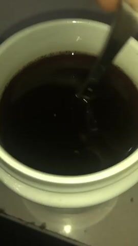 A close-up overhead shot shows a white mug filled with dark liquid, presumably coffee, which is being stirred with a metal spoon. The liquid has a thin layer of foam around the rim. The background is dark and out of focus. The spoon is positioned in the upper right quadrant of the mug.