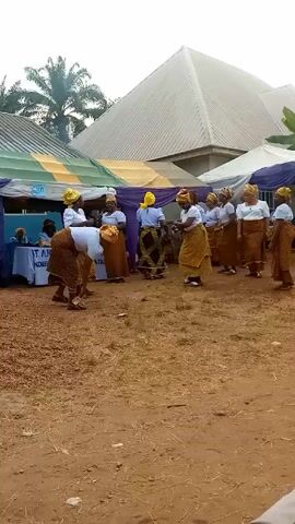 The video shows a group of women dancing in a circle outdoors. They are dressed in matching outfits consisting of white tops, patterned brown and gold skirts, and yellow head wraps. The ground is dirt, and there are scattered brown objects, possibly seeds or nuts. In the background, there are tents and a building with a light-colored roof. A sign is visible, reading "ST. ANTHONY OF NDIEZIKIE, ANAMBRA ST." There appear to be people seated under the tents. The women are actively moving and seem to be engaged in a traditional dance. The overall setting suggests a cultural event or celebration. The perspective is from a static camera position.