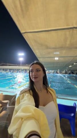 The video shows a young woman in a yellow jacket and white top posing for a selfie. She is standing under a covered area at night, with a swimming pool behind her. Several lanes are visible in the pool, with people swimming in them. There are lights illuminating the pool area. The woman smiles and turns her head slightly as she records the video.
