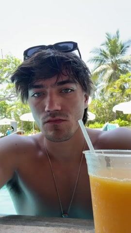 A light-skinned man with dark hair drinks from a straw, in what appears to be an orange juice beverage in a plastic cup. The man has sunglasses on his head, and is wearing a silver chain. Palm trees, beach umbrellas, and people can be seen in the background. The man appears to be at a resort or in a tropical location.