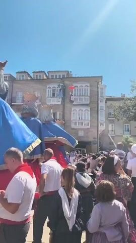 This is a vertical, medium shot video taken outdoors on a sunny day. The scene is crowded with people of various ages, many of whom are looking towards the left of the frame. Two very large, brightly colored figures, appearing to be giant puppets or effigies of kings with crowns, dominate the left side of the image. They are dressed in blue cloaks with red and gold trim. Between them, a bright red balloon or object is visible, suspended in the air. In the background, a multi-story building with balconies is visible, and people are gathered on some of the balconies. A person can be seen hanging from a wire attached to one of the balconies. The crowd is dense, suggesting a festive or celebratory event, possibly a parade or festival. The people in the foreground are dressed in casual clothing, with some wearing traditional attire or accessories, like red scarves. The overall atmosphere appears to be lively and engaging.