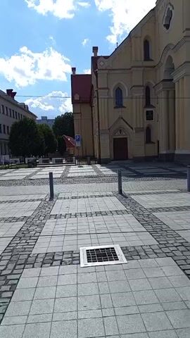 The video pans across a paved public square. A large, light yellow building with a red roof, which appears to be a church or cathedral, dominates the right side of the frame. The building features arched windows and columns. In the foreground, the square is paved with alternating squares of light and dark gray tiles, with decorative bands of smaller dark cobblestones. Two metal bollards are visible in the square, along with a square metal grate in the pavement. To the left of the church, a modern multi-story building with blue and gray accents is visible. A few people are walking in the distance near this building. The sky above is blue with scattered white clouds. The scene appears to be a bright, sunny day.