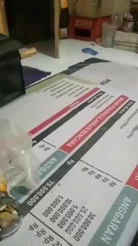 The video shows a close-up of a person's legs and feet lying on a white banner with text and financial figures printed on it. The banner appears to be on a floor. The text on the banner is in Indonesian and includes the words "ANGGARAN" (budget) and "PEMBANGUNAN MASYARAKAT" (community development). There are also monetary values like "Rp. 75.500.000". In the background, there are some objects on the floor, including a plastic bag, an ashtray with cigarette butts, and some boxes. A piece of furniture, possibly a cabinet, is visible in the upper right corner. The lighting is dim, suggesting an indoor setting, possibly a room.