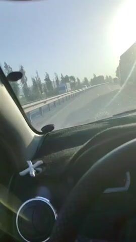 The video shows a first-person perspective from inside a car driving on a highway. The sun is bright and overhead, casting a glare on the windshield. A white, star-shaped air freshener is visible on the dashboard. The side mirror shows other vehicles on the highway, including a white truck. Trees and hills are visible on the side of the road. The driver's side door panel is also visible.