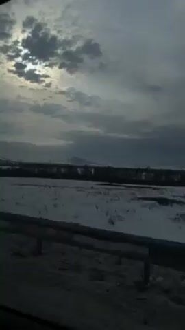 The video shows a view from a moving vehicle, likely a car, looking out at a snowy landscape. The foreground includes a portion of the vehicle's exterior, specifically the roadside barrier. Beyond that is a field covered in snow, with sparse vegetation visible. The sky is overcast with dark clouds partially obscuring the sun, which is visible as a bright spot. In the distance, there are hints of a skyline or mountain range, but the visibility is limited due to the weather conditions. The overall tone is muted and somewhat bleak due to the gray sky and snow-covered ground.