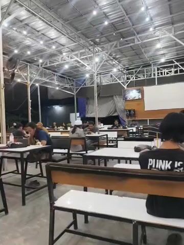 The video shows the interior of a cafe or restaurant with multiple tables and chairs. The space is covered by a metal roof structure adorned with numerous string lights. Several patrons are seated at tables, some appearing to be eating or engaged in conversation. In the foreground, a person is seated with their back to the camera, wearing a black t-shirt with "PUNK IS" visible on the back. The overall atmosphere appears casual and relaxed, with diners scattered throughout the venue. The flooring is a smooth, light-colored concrete or stone.