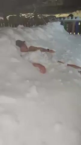 A man lies nearly submerged in a snowdrift, wearing a hat. His torso, arms, and legs are visible. He is rubbing snow onto his bare skin.  A wooden fence borders the snowdrift.