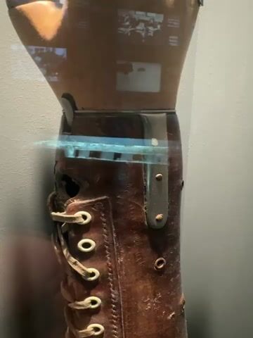 A close-up, medium shot shows a prosthetic leg made of brown leather. The leg has laces running up the side, and metal eyelets. There are also metal straps and rivets visible on the leg. The prosthetic is likely on display in a museum or exhibit, as there appears to be a glass case in front of it, reflecting the surrounding environment, including what looks like a projected image or video on the upper part of the prosthetic. The background is a plain gray wall. The lighting is dim, with a blueish-green light effect across the middle of the prosthetic.