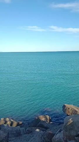 The video shows a static shot of the ocean taken from a rocky shoreline. The water is a vivid turquoise blue, gently rippling, extending to a distant horizon where a faint landmass can be seen. The sky is a clear, bright blue with a few sparse, wispy clouds near the top of the frame. The foreground is filled with gray and brown rocks of various sizes, creating a natural barrier between the viewer and the water's edge.