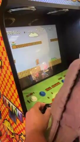 A man is playing Super Mario Bros on a multi-game arcade machine. The machine is adorned with artwork of other arcade classics like Pac-Man and Tetris. The game displays Mario jumping through the Mushroom Kingdom. The man operates the joystick with his left hand.