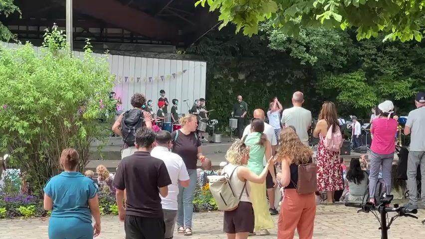 The video shows a group of people gathered outdoors, seemingly at an event or concert. In the background, a band is performing on a stage. Some band members are playing instruments, while others appear to be singing or interacting. A small crowd is visible watching the performance. In the foreground and mid-ground, more people are standing and observing. The environment appears to be a park or an outdoor venue with trees and greenery surrounding the stage area. The overall atmosphere seems casual and festive.
