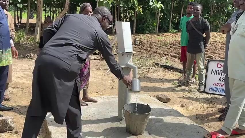 The video shows a group of people gathered around a manual water pump in an outdoor setting. A man in a black outfit is operating the pump, holding a metal cup under the spout. A woman in a patterned skirt and striped shirt is assisting with the pump's handle. Water is visibly flowing into the cup. Another person, a man in a blue shirt, is seen to the left, and several other people, mostly men, are standing around observing the activity. In the background, there are trees and vegetation, suggesting a rural or semi-rural environment. A sign with text is partially visible on the right side of the frame. The ground is dirt and concrete.