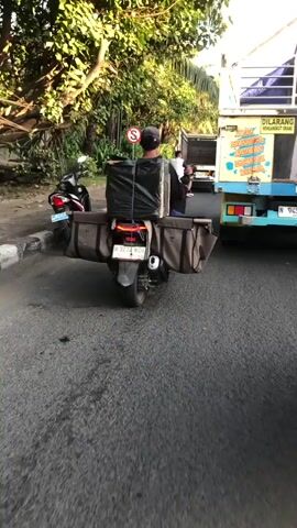 The media shows a street scene in Klurak, Indonesia. The primary subject is a person riding a scooter with a large, black, plastic-wrapped package strapped to the back. The package appears to be secured with ropes and is quite tall. The scooter also has two large, canvas-like bags attached to its sides, extending outwards. The rider is wearing a dark baseball cap and a dark shirt, with their back to the camera.

Behind the scooter, a large yellow truck is visible, also on the road. Part of the truck's side is visible, featuring some text. The truck appears to be stopped or moving slowly, as is the scooter. Further behind the truck, another scooter with a rider can be seen. The scene appears to be a typical street with trees and foliage lining one side, suggesting an outdoor, possibly tropical setting.

The lighting suggests it is daytime, likely late afternoon given the angle of the sun casting shadows. The weather appears to be clear and fair. The road is paved, and there is a curb on the left side of the image. A red and white "no U-turn" sign is visible on a pole on the left side of the road.

Visible text on the truck includes: "DILARANG", "MEMBUANG SAMPAH SEMBARANGAN", and "MENGANGKUT URANG". The license plate on the scooter reads "W 5274 NGH". The overall impression is a snapshot of daily life and transportation on a street in Indonesia, highlighting a method of carrying goods via scooter.