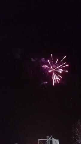 The video captures a fireworks display at night. The sky is dark, with the fireworks creating bursts of light and color. One prominent firework is a large, multi-colored explosion with radiating sparks. Another firework is a smaller, red projectile ascending. A green firework is also visible, ascending and emitting sparks. In the lower part of the frame, structures that appear to be part of a stage or scaffolding are present, and the bottom edge of some other fireworks are visible. The audio includes cheers and commentary, suggesting an audience is present.