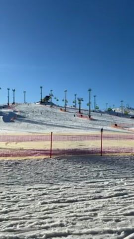 The video shows a ski slope on a clear, sunny day. The sky is a bright, solid blue. The ground is covered in snow, with visible tracks from skiers or snowboarders. A yellow and red safety fence runs across the lower part of the frame. Further up the slope, there are several ski lifts with chairs visible, and tall light poles scattered across the hillside. A few people can be seen skiing or snowboarding down the slope, appearing as small figures in the distance.