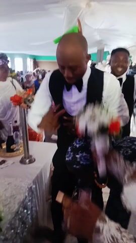 Here's a description of the video:

The video shows a wedding celebration in a decorated indoor space. A bald man wearing a white shirt, black vest, and bow tie is dancing. Another man, also in a similar outfit, stands behind him, smiling. In the foreground, a woman in a white dress interacts with another woman wearing a patterned blue headwrap and top. Other guests are visible in the background, some taking pictures. The setting appears to be a reception area, with tables and decorations.