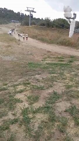 A group of people are running up a grassy, dusty hill. In the background, there is a ski lift with several gondolas. The sky is cloudy. The people are wearing casual clothing, and some are wearing athletic shoes. It appears to be a race or a recreational activity. The video is shot from a low angle, looking up the hill.