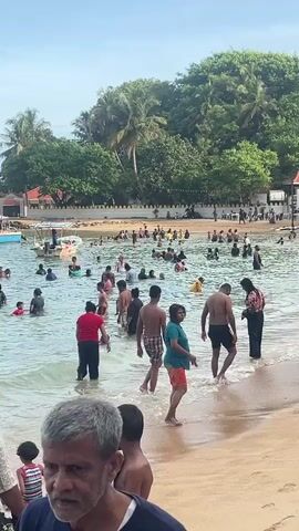 The video shows a crowded beach with many people swimming and wading in the water. The water is calm and clear. There are a few boats docked in the distance. The beach is sandy and there are trees and buildings in the background. Several people are standing on the shoreline watching the swimmers. There are some people with dark skin.  The sky is blue and the weather appears to be sunny.