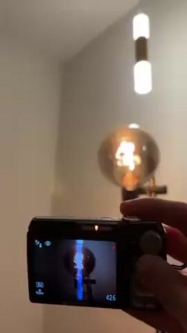 A person holds a black Olympus digital camera, pointing it at a room with two light sources. The camera's screen displays a blue-tinged image of a large, round, old-fashioned incandescent light bulb on a stand, which is also visible in the room. Above it, a modern, vertical, cylindrical light fixture with two white bulbs and a brown middle section is hanging from the ceiling. The walls are a neutral, light color. The camera is slightly shaky, and the person is likely taking a picture of the light bulbs.