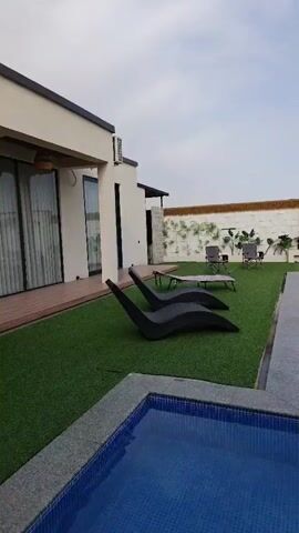 This is a video of an outdoor swimming pool and surrounding area. In the foreground, there is a blue tiled swimming pool. To the left of the pool, there is artificial turf with two black lounge chairs. In the background, there is a white wall with some decorative elements, and a building with a modern design. There is also a palm tree and a small covered seating area with chairs and a table. The sky is overcast.