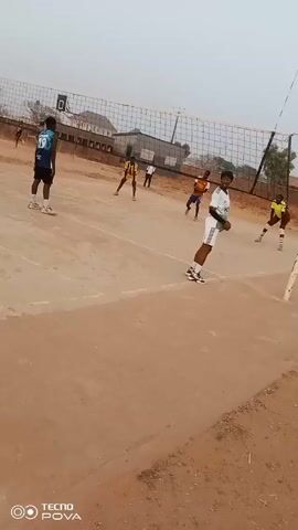 The video shows a volleyball game taking place on an outdoor court. Several players are positioned on either side of the net, ready to play. The court appears to be a hard surface, possibly concrete or asphalt, with visible lines marking the boundaries. The net is stretched between two poles. In the background, there are buildings and a fence visible. The sky is overcast, suggesting it might be a cloudy day. The video appears to have been taken with a TECNO POVA phone.