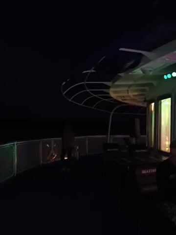 The video shows a dark outdoor setting, possibly a deck of a boat at night. There are railings with white netting and blue accents. Colorful lights are projected onto a person standing near the railing. The person is wearing a dark outfit and appears to be interacting with the lights. Another person is partially visible in the background. The overall atmosphere is dimly lit with dynamic lighting effects.